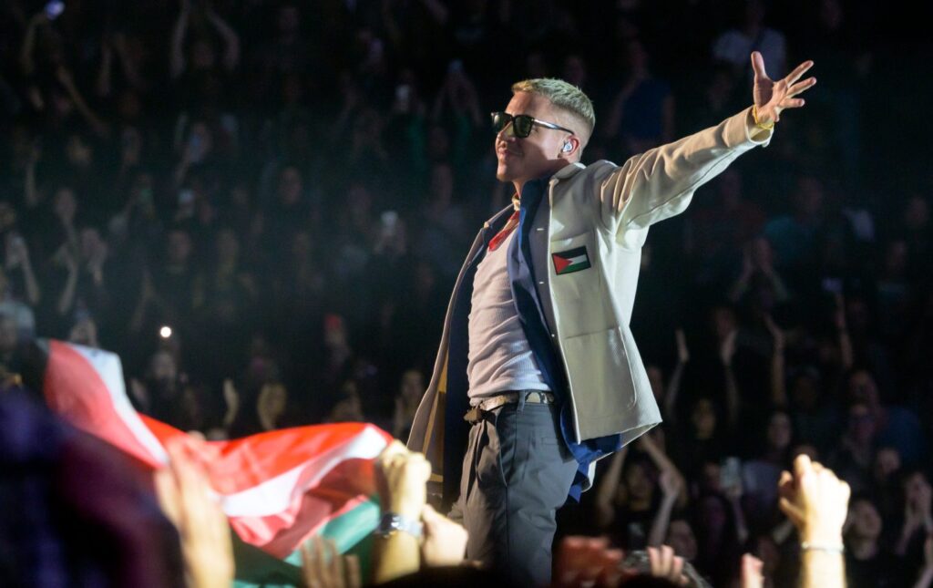 Macklemore, wearing sunglasses and a jacket emblazoned with a Palestinian flag, gestures to the crowd at the concert.